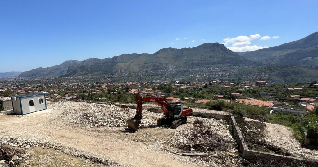 Il lotto di via Ignazio Florio a Monreale visto dalla strada, con una cabina di cantiere, un escavatore e i monti sullo sfondo